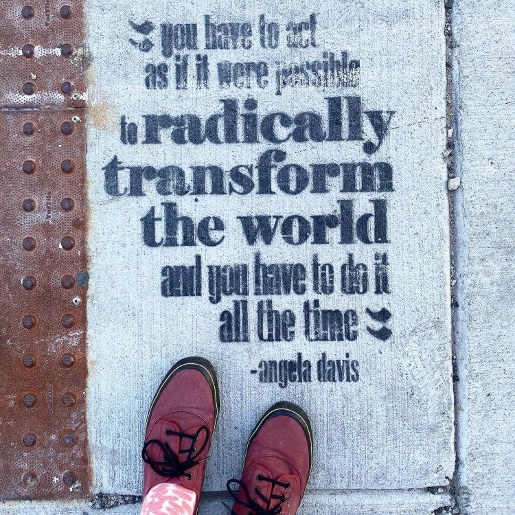 Lee's shoes on the ground below a spray painted stenciled quote: you have to act as if it were possible to radically transform the world and you have to do it all the time -angela davis