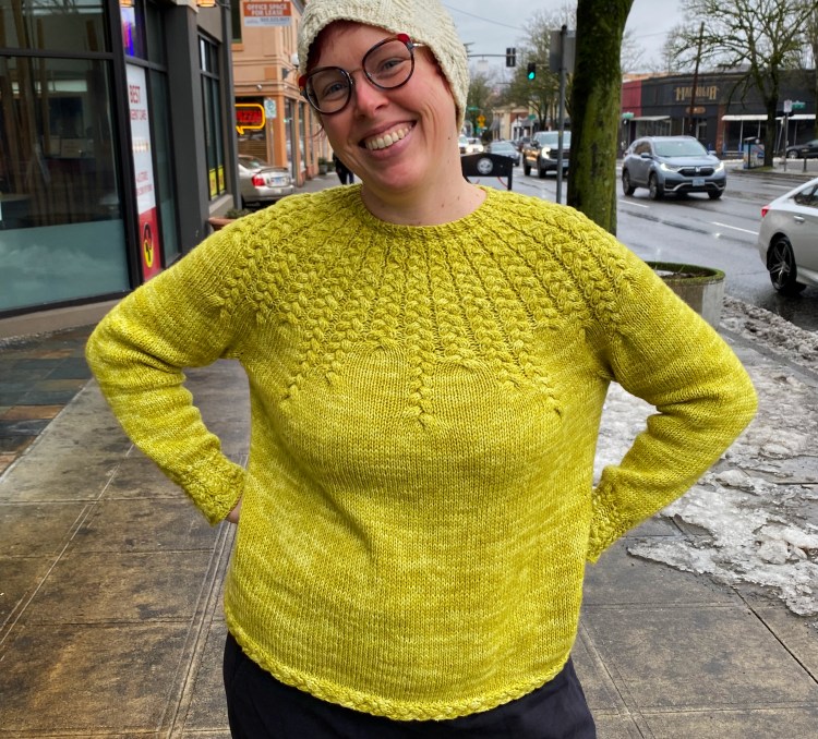 Lee wearing the sweater, looking forward and smiling, standing on the sidewalk, hands on hips