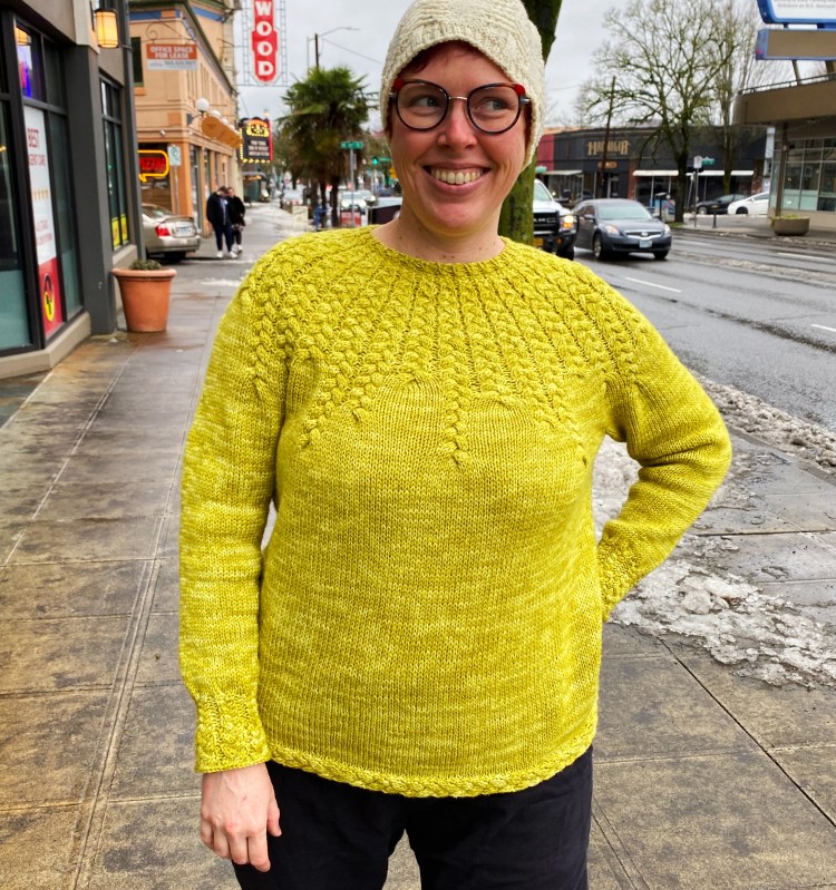 Lee wearing the sweater, looking forward and smiling, standing on the sidewalk
