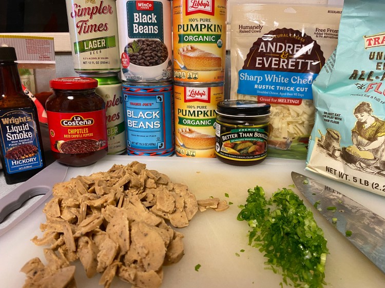 ingredients: a cutting board with fake sausage sliced thinly and green onions with a knife; liquid smoke, chipotles, simpler times lager, black beans cans, pumpkin cans, better than bouillon vegetable base, sharp white cheddar, all-purpose flour