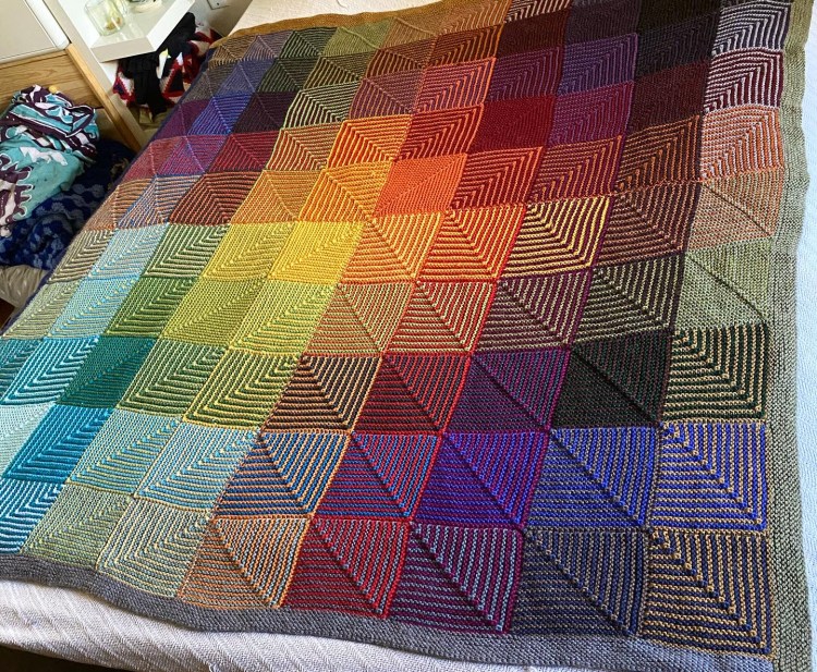 Hue Shift rainbow colored mitered square blanket laying flat on a bed