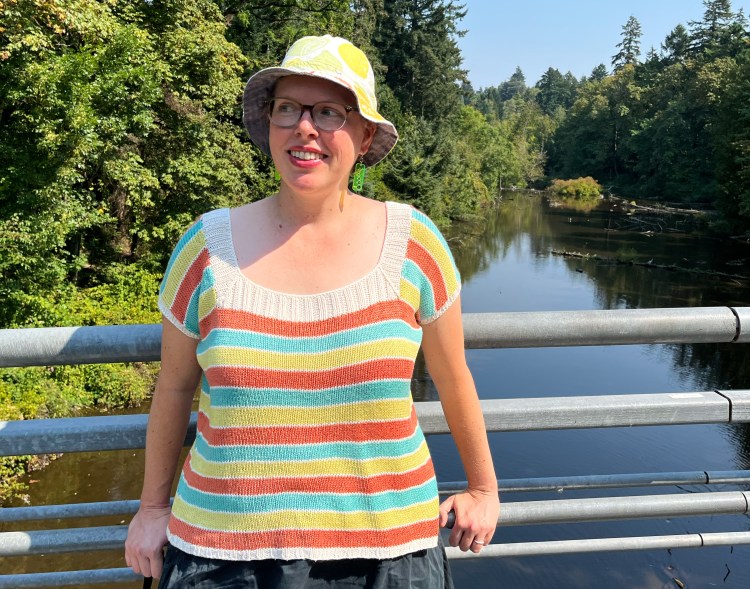 Lee standing on a bridge with a river behind, wearing a green/orange/aqua striped knit tee and a white and yellow/green bucket hat