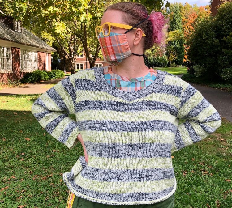 Lee looking to the side wearing a plaid mask that matches the top that's peeking out under her striped speckly green and black cotton knit sweater