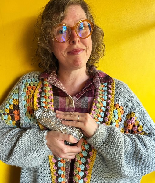 Lee holding a log of seitan, wearing a crocheted cardigan over a flannel and round glasses