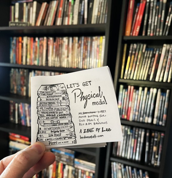 hand holding up small "Let's Get Physical Media!" zine with subtitle "My [addiction, crossed out] hobby movie hunting for dvd deals & blu ray bargains. a zine by Lee leedoesstuff.com, with dvds in the background