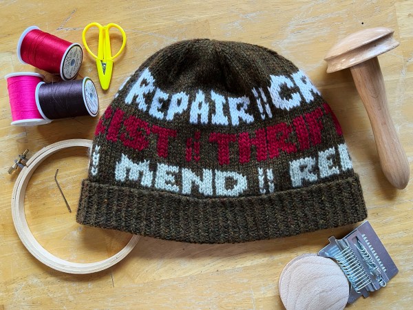hat with words on it laying flat amongst related items (darning mushroom and loom, thread, embroidery hoop)—visible words are REPAIR, THRIFT, MEND