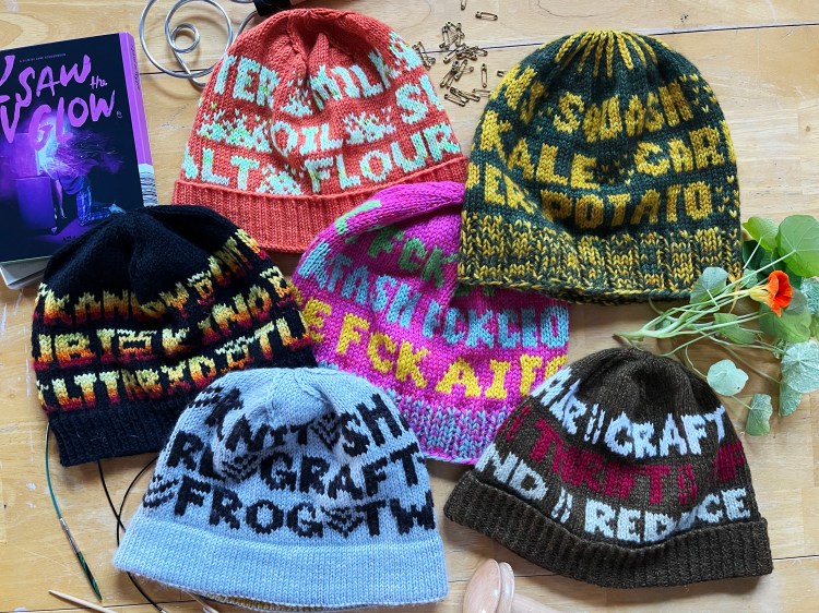6 hats with words on them laying flat amongst related items (blu ray, safety pins, nasturtiums, darning mushroom, knitting needles)—visible words include OIL, FLOUR, SQUASH, KALE, POTATO, LTTRBXD, FCK AI, GRAFT, FROG, CRAFT, THRIFT, REDUCE