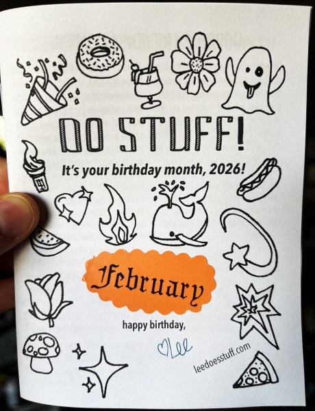 hand holding zine that says "Do Stuff! It's your birthday month, 2026!" with drawings of emojis and orange cloud-shaped sticker that says "February"